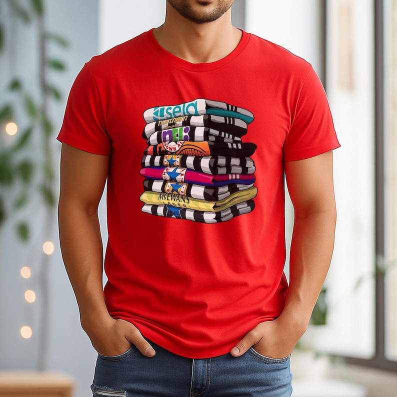 May include: A red t-shirt featuring a stack of colorful, folded items with text. The items are in various colors, including blue, black, yellow, and pink. The text on the items includes the word "ISELD" and "MCEWANS".