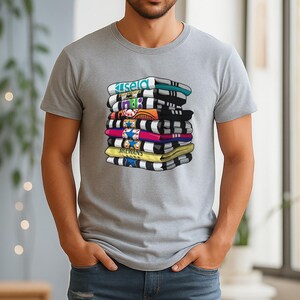 May include: A gray t-shirt featuring a graphic of a stack of folded towels with various designs and colors. The towels have text that reads "Kisela", "MTB", and "MCEWANS". The towels are in shades of blue, black, yellow, pink, and orange.