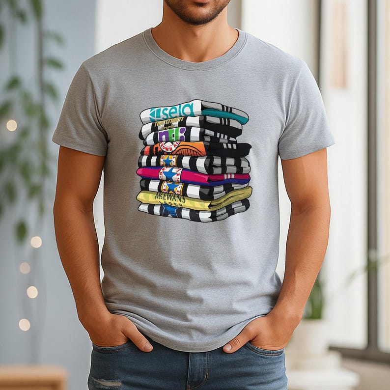 May include: A gray t-shirt featuring a graphic of a stack of folded towels with various designs and colors. The towels have text that reads "Kisela", "MTB", and "MCEWANS". The towels are in shades of blue, black, yellow, pink, and orange.