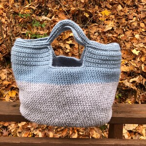 May include: A crocheted tote bag with a light blue and gray color scheme. The bag has a wide, open top and two handles.