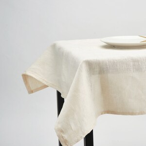 May include: A white linen tablecloth draped over a table with a white plate and a gold fork on top.