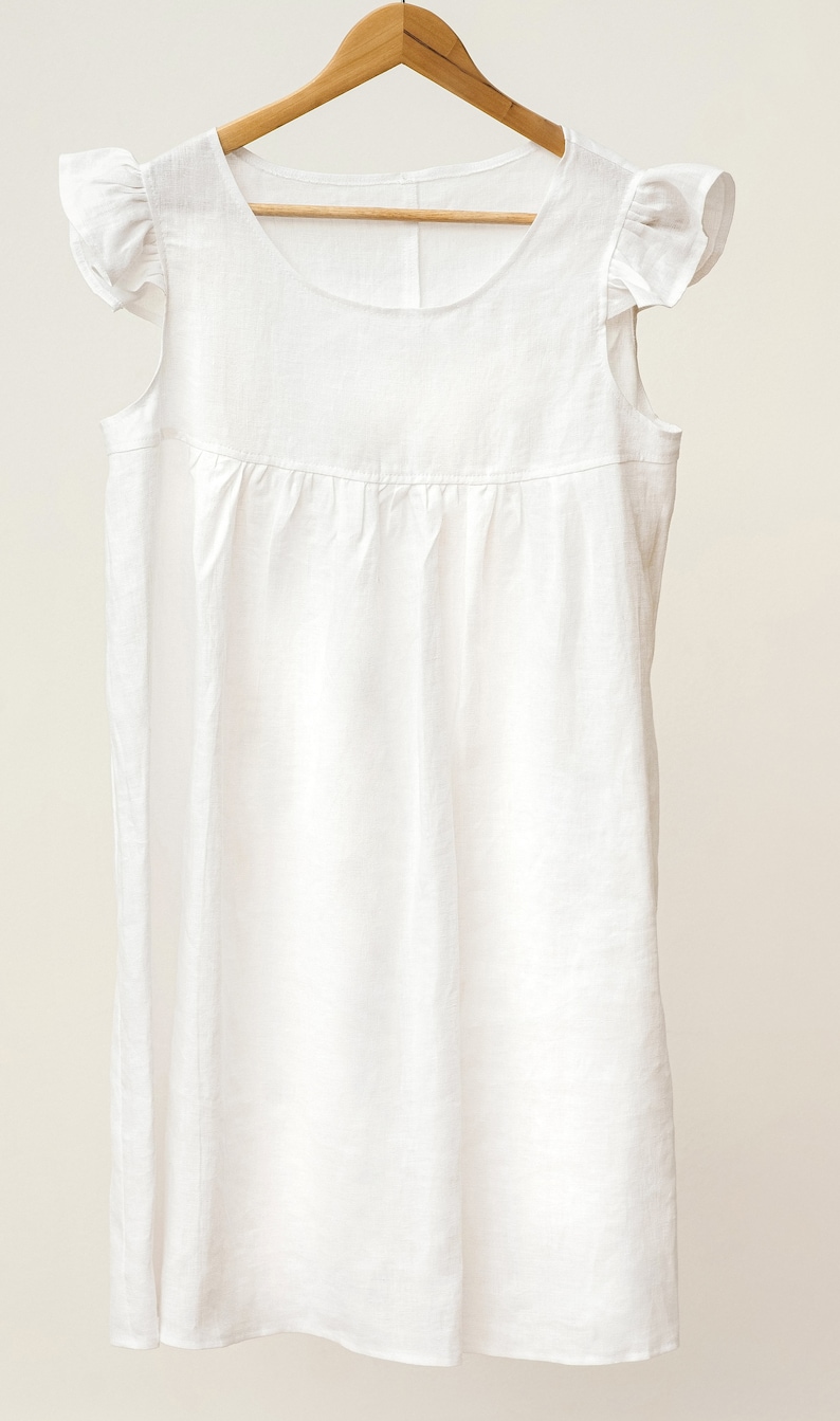 Puede incluir: Un vestido blanco de lino con mangas de volantes. El vestido tiene un escote redondo y una falda fruncida.