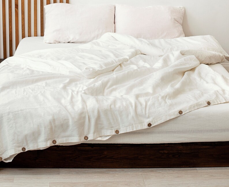 Op de afbeelding: Een wit linnen dekbedovertrek met houten knopen op een bed met een houten hoofdeinde. Het dekbedovertrek is licht gekreukt en het bed is opgemaakt met witte lakens en kussens.