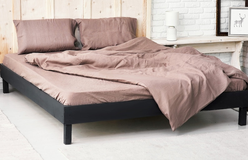 Op de afbeelding: Een bruin linnen bedovertrek met twee bijpassende kussenslopen op een zwart houten bedframe. Het bed is gemaakt met een bruin linnen dekbedovertrek en het bedframe heeft een eenvoudig ontwerp.