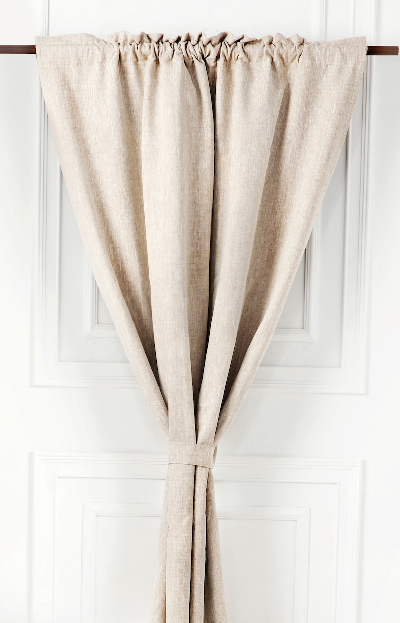 May include: A beige linen curtain with a gathered top and a tied bottom, hanging from a rod against a white wall.