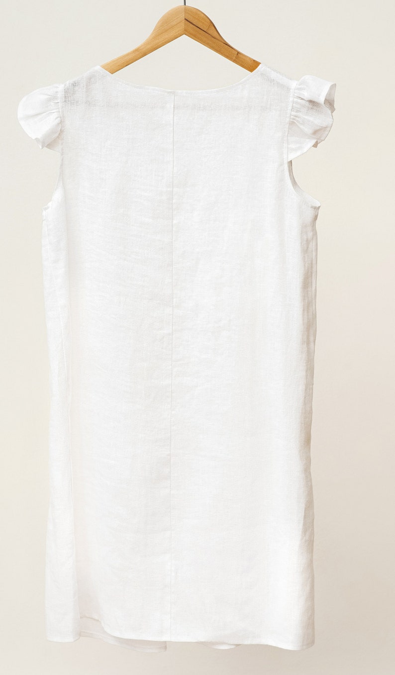 Puede incluir: Vestido blanco de lino con mangas de volantes. El vestido tiene un escote redondo y un corte holgado.