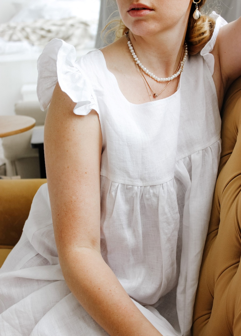Puede incluir: Un vestido blanco de lino con mangas con volantes y un escote cuadrado. El vestido lo lleva una persona con un collar de perlas y un collar de cadena dorada.