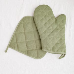May include: A set of two green quilted oven mitts. The mitts are made of a soft, textured fabric and have a diamond pattern.