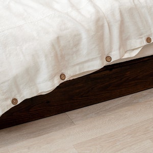 Op de afbeelding: Een wit linnen beddengoed met houten knopen langs de rand, gestopt in een donkerhouten bedframe.