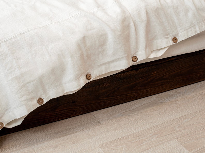 Op de afbeelding: Een wit linnen beddengoed met houten knopen langs de rand, gestopt in een donkerhouten bedframe.