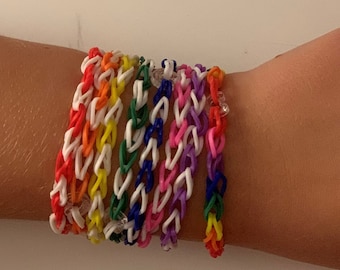 May include: A stack of colorful rainbow loom bracelets on a person's wrist. The bracelets are made of rubber bands and are in a variety of colors, including red, orange, yellow, green, blue, and purple.
