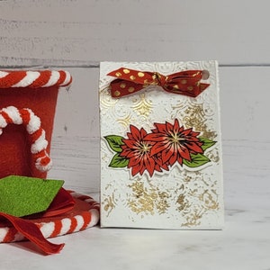 May include: A white gift bag with a red and white polka dot bow and a red poinsettia design. The bag has a gold glitter background.