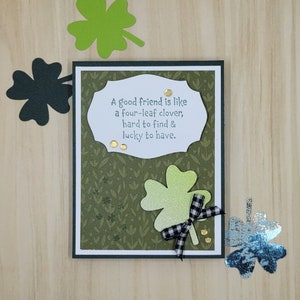 May include: A green card with a white border and a white rectangle in the centre. The rectangle contains the text "A good friend is like a four-leaf clover, hard to find & lucky to have." There are three gold glitter dots below the text. A green four-leaf clover with a black and white checked bow is on the card.