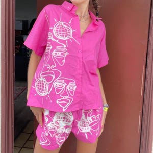 May include: A pink button-up shirt and matching shorts set with a white abstract face and sun design.
