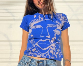 May include: A blue t-shirt with a white abstract face design. The shirt has a crew neck and short sleeves.