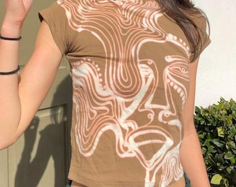 May include: A brown short-sleeved t-shirt with a white abstract design of a face. The design is made up of swirling lines and dots.