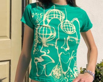 Könnte beinhalten: Grünes T-Shirt mit kurzen Ärmeln und einem weißen Grafikdruck von zwei abstrakten Gesichtern mit geometrischen Mustern.