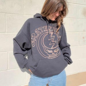 May include: A dark gray hooded sweatshirt with a brown graphic of the Grateful Dead logo. The logo features a skull with a lightning bolt and the words "Grateful Dead" in a circle around it.