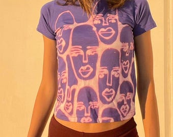 May include: A purple t-shirt with a pattern of pink outlined faces. The faces are abstract and have no eyes, but have noses and mouths.