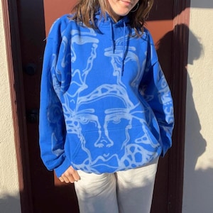 May include: A royal blue hoodie with a light blue abstract face design. The hoodie has a front pocket and a drawstring hood. The person is wearing white pants. The design is repeated across the front and sleeves of the hoodie.