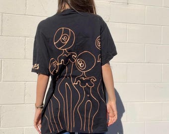 Könnte beinhalten: Schwarzes Oversize-T-Shirt mit einem grafischen Druck von drei Figuren mit 8-Ball- und 10-Ball-Designs. Die Figuren sind in hellbrauner Farbe umrandet.