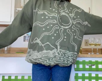 Könnte beinhalten: Ein grüner Kapuzenpullover mit einem weißen Sonnen- und Wellendesign. Der Pullover ist aus einem weichen, bequemen Material gefertigt und hat eine lockere Passform.