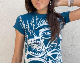 May include: Teal short-sleeve t-shirt with a white abstract design. The graphic includes stars, swirls, and geometric shapes. The shirt has a crew neck and a fitted silhouette, suitable for casual wear.
