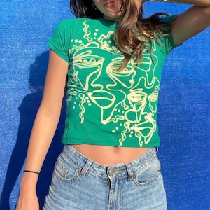May include: Green cropped t-shirt with a white abstract face design. The shirt is made of a soft, comfortable material.