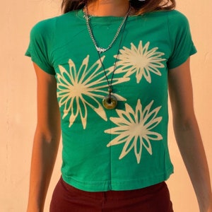 May include: A green t-shirt with a white floral print. The shirt has a crew neck and short sleeves. The model is wearing a necklace with a green stone pendant.