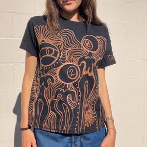 May include: Black t-shirt with a brown abstract design featuring a swirl pattern, figures, and the numbers "2" and "8".