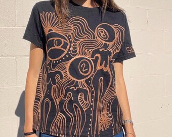 May include: Black t-shirt with a brown abstract design featuring a swirl pattern, figures, and the numbers "2" and "8".