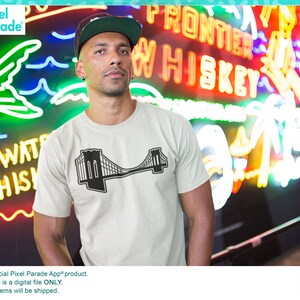 May include: A beige t-shirt with a black graphic of the Brooklyn Bridge. The t-shirt is being worn by a person standing in front of a neon sign that says "Frontier Whiskey".
