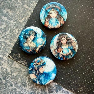 May include: Four circular buttons with illustrations of women in blue and white, holding orbs. The women have long hair and floral headpieces. The buttons have a mystical, fantasy theme and are set against a dark background.