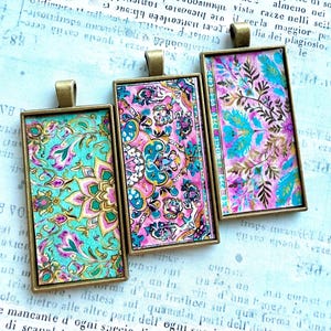 May include: Three rectangular pendants with ornate, colorful patterns in teal, pink, and gold. Each pendant has a gold-colored frame and a loop at the top for a chain. The designs feature floral and paisley motifs.