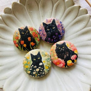 May include: Four round cat-themed decorative pins. Each pin features a black cat with yellow eyes, set against a backdrop of colorful flowers. The pins are arranged on a white, petal-shaped dish, with dried plant stems in the background.