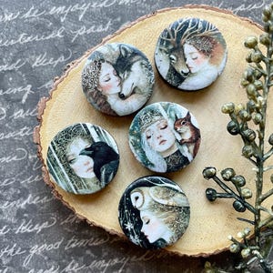 May include: A collection of five round, decorative pins featuring fantasy art. Each pin showcases a woman with an animal, including a wolf, deer, raven, squirrel, and owl. The pins are arranged on a wooden slice.