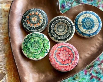 May include: Five round wooden coasters with intricate mandala designs. The coasters feature different colors and patterns, including black, white, green, blue, and pink.