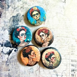May include: Five circular pins featuring artistic portraits of a person with dark hair adorned with red flowers. Each pin displays a unique colour palette, including shades of blue, orange, and sepia, with detailed facial features and decorative elements.