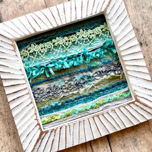 May include: A framed artwork featuring a layered textile design with various shades of green, blue, and grey. The frame is white with a textured, raised edge.