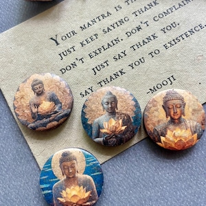 May include: Four round Buddha image pins with a vintage aesthetic. Each pin features a different Buddha image holding a lotus flower. A card with the text "Your mantra is thank you" is in the background.
