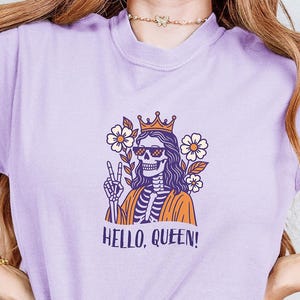 May include: Orchid-colored t-shirt featuring a skeleton wearing sunglasses and a crown, with the text "HELLO, QUEEN!". The graphic includes flowers and a peace sign. The shirt is tied at the waist and paired with blue jeans.