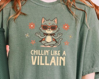 May include: Olive green t-shirt with a graphic of a meditating cat wearing sunglasses, surrounded by flowers, and the text "CHILLIN' LIKE A VILLAIN". The shirt is a relaxed fit.