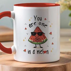 Op de afbeelding: Witte keramische mok met een rode binnenkant en handvat. De mok heeft een watermeloenpartje met een zonnebril op en maakt een hartvorm met zijn handen. De tekst op de mok luidt "You are one in a melon."