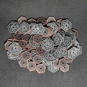 Puede incluir: Un montón de fichas de juego hexagonales en tonos grises y cobrizos. Cada ficha presenta un símbolo de átomo y el número "5". Las fichas están dispersas sobre una superficie gris oscuro.