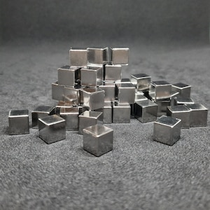 May include: A pile of small, silver-coloured metal cubes. The cubes are arranged in a pyramid shape, with some cubes scattered around the base.