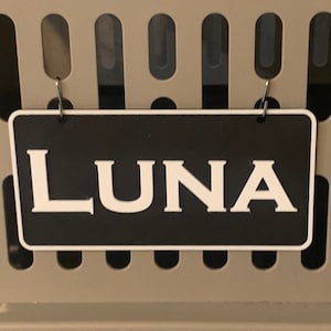 May include: A beige pet carrier with a black metal grate. A black and white sign with the name "LUNA" is attached to the front of the carrier.
