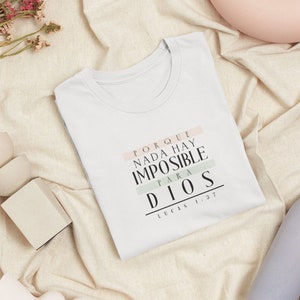 May include: White t-shirt with a quote in Spanish that reads "PORQUE NADA HAY IMPOSIBLE PARA DIOS LUCAS 1:37". The quote is printed in black with pink and green stripes.