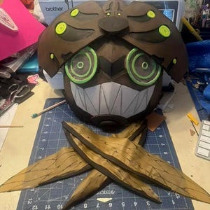 May include: A brown and black mask with green and white accents, featuring a stylized face with large eyes and a toothy grin. The mask is paired with two golden-brown wing-like structures. A sewing machine is visible in the background.