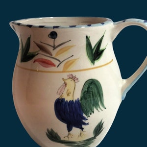 May include: A white ceramic pitcher with a blue, green, yellow, and pink rooster design. The pitcher has a handle and a wide mouth.