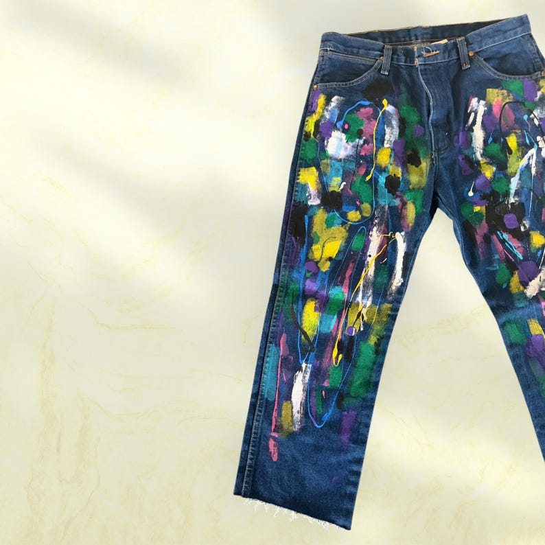 Pu&ograve; includere: Jeans in denim blu scuro con un colorato design astratto a spruzzi di vernice. La vernice comprende bianco, giallo, viola, verde e blu. I jeans hanno un classico design a cinque tasche e un orlo grezzo.